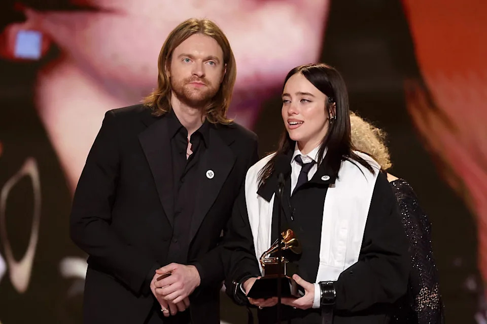 Two individuals stand on stage at an awards event. One holds a trophy. Both are dressed in formal attire, including a suit and a jacket with a tie