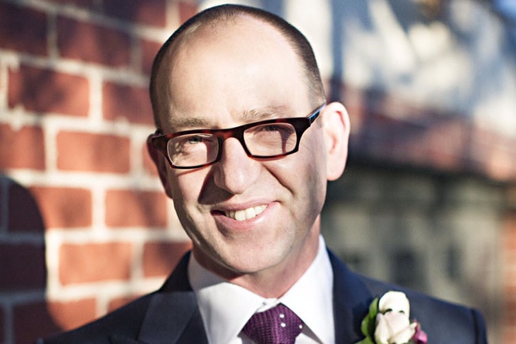 A portrait of a man smiling at the camera. He his standing outdoors wearing a suit and tie and has a flower in his lapel.