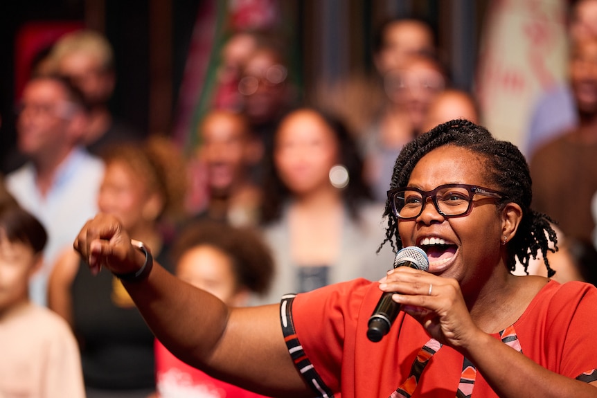 Buyi Zama, a Black woman holding a mic, singing, arm raised. Blurred cast behind her.
