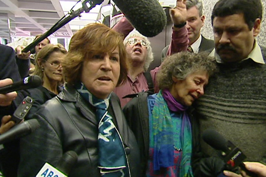 Two women stand in front of microphones and a media pack. One woman leans on a man for support. 