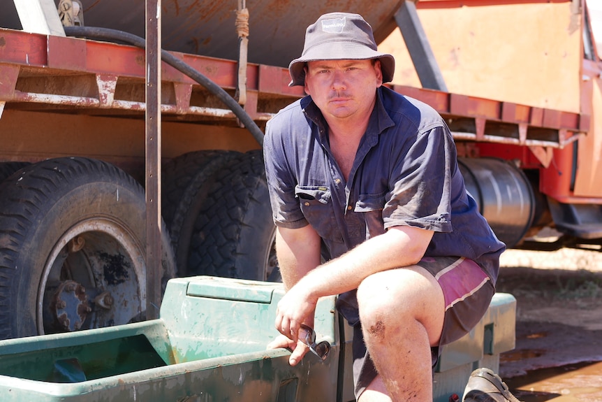 A man in a blue shirt and hat kneels near a water trough