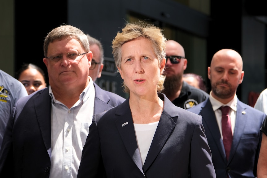 A woman in a suit talking to the press flacked by men in suits. 