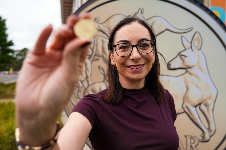 A woman with dark brown hair and glasses holds up a gold coin, in front of a large poster of a coin.