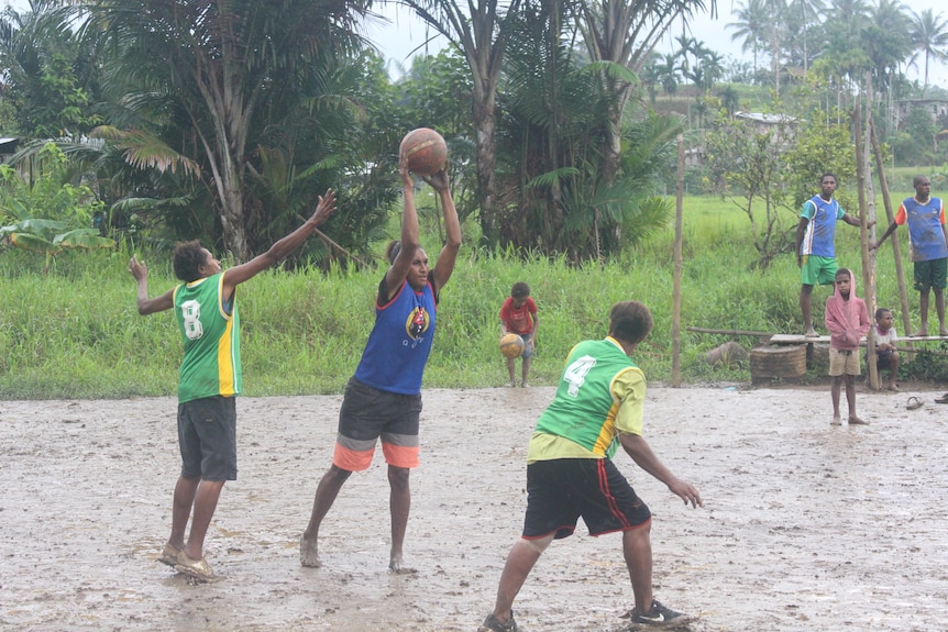 Three people play basketball on a muddy court in bare feet.  The lady in the middle holds the basketball to pass it