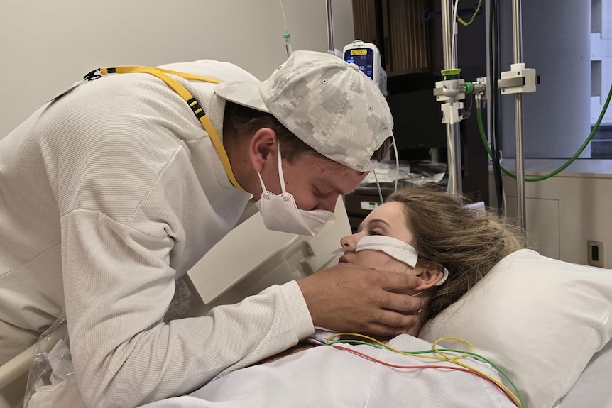 A man with a face mask and backwards facing cap touches the face of a young woman in a hospital bed.