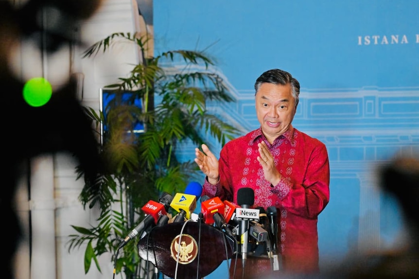 A man in a red shirt speaks at a podium with a microphone.