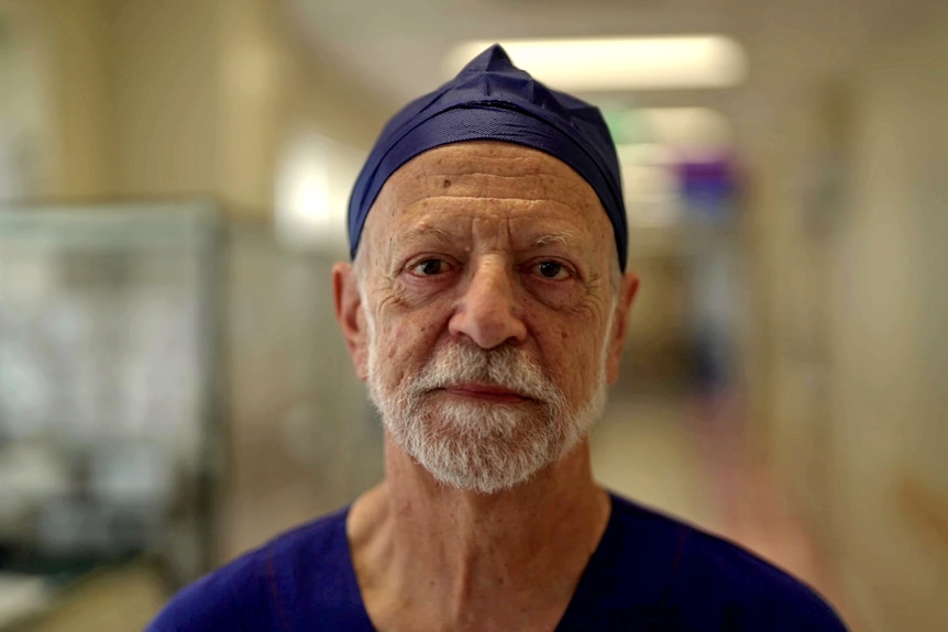 An older man with a short grey beard wears a navy surgical scrub cap on his head standing in a hospital corridor.