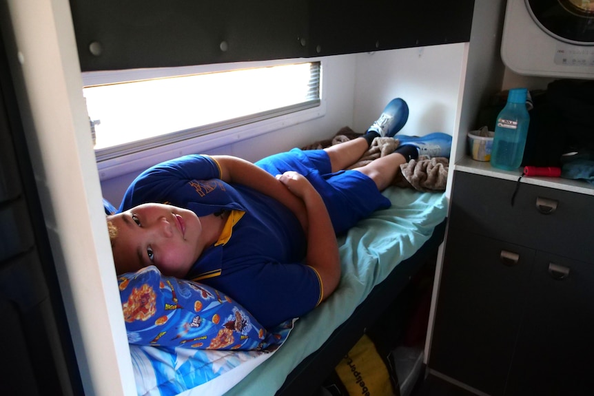 A boy in a school uniform lies on a small bunk in a caravan.