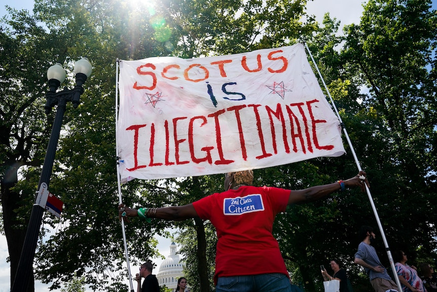 Protester holds a sign that reads "SCOTUS is illegitimate" while wearing a red T-shirt with text that reads "2nd Class Citizen"