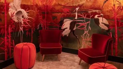 Tropical birds and plants on the walls of the washroom with red and yellow tropical scenes across the walls. There are red velvet chairs on the ground.