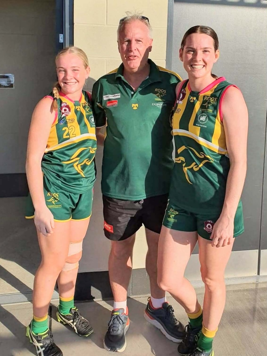 Two smiling young women in Aussie Rules uniforms with an older man in a matching polo shirt.