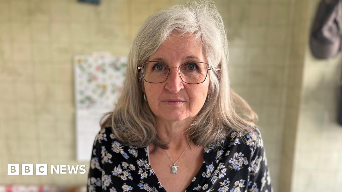 A woman with grey hair, glasses and a dark blue blouse with flowers is looking into the camera. She is standing in her kitchen.