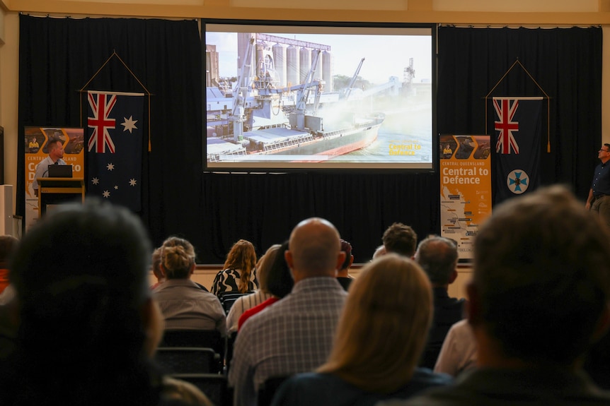 A powerpoint with flags and signs next to it, with people watching in a dimly lit room