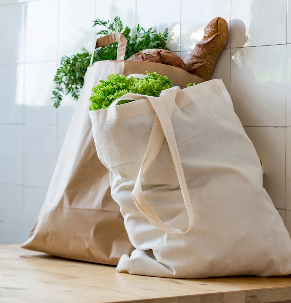 Two reusable shopping bags on a countertop, filled with fresh vegetables and baguettes, against a tiled wall