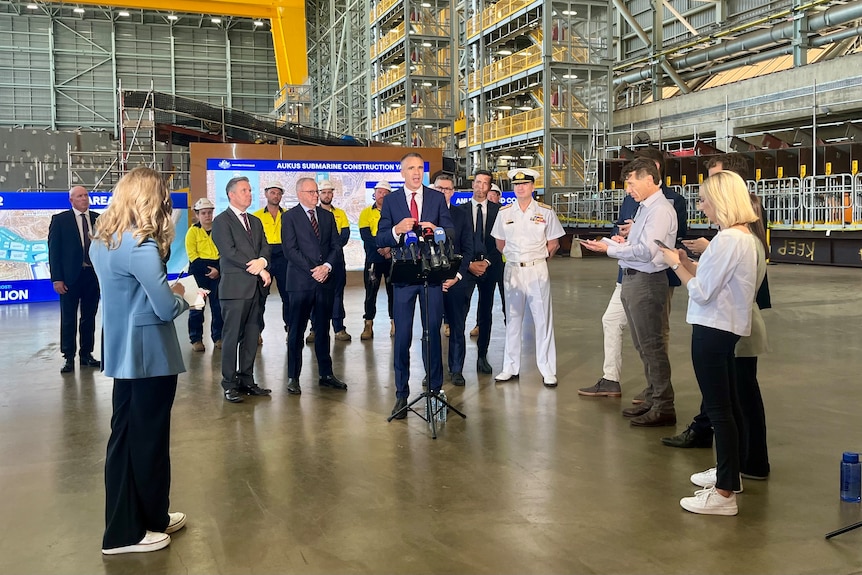A man wearing a suit and tie standing in front of others speaks to reporters in a large industrial building