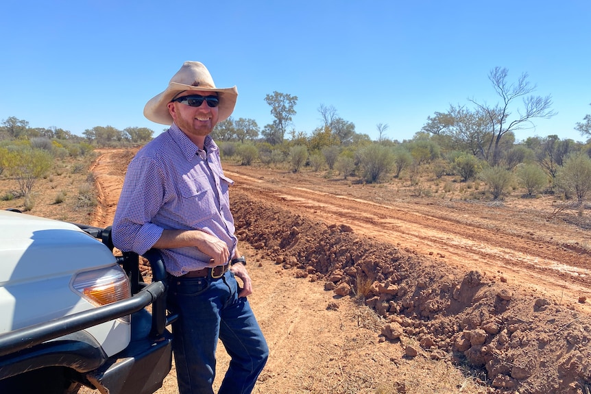A man leans on a bullbar of car surrounded by red dirt.