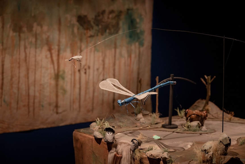 In a dark gallery space sits a miniature set, which includes a model of a mosquito. It flies above a waterfall with eyes.
