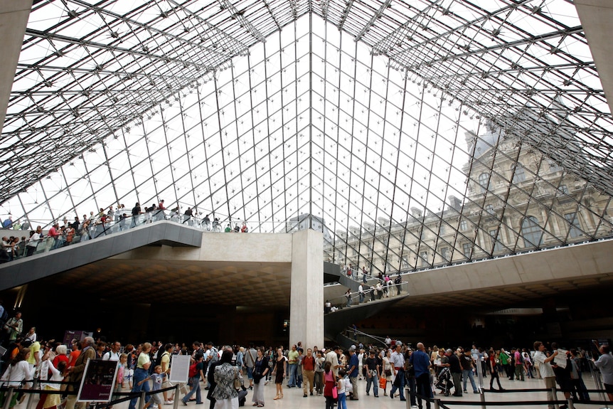 Groups of people walking in a mass underneath the gridded glass pyramid roof of the Louvre lobby.