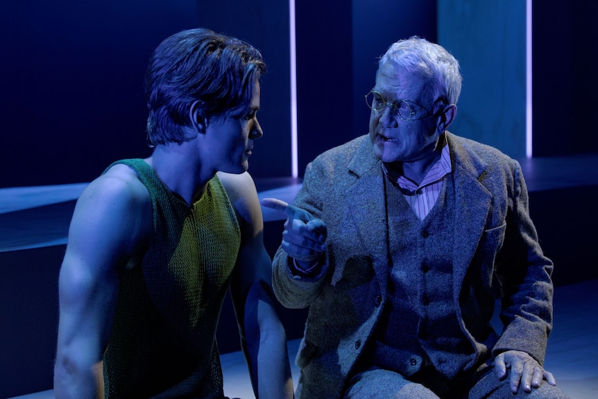 On stage, bathed in blue light, sits Simon Burke, early 60s, in a three-piece suit, waggling a finger at Tom Rodgers, mid-20s..