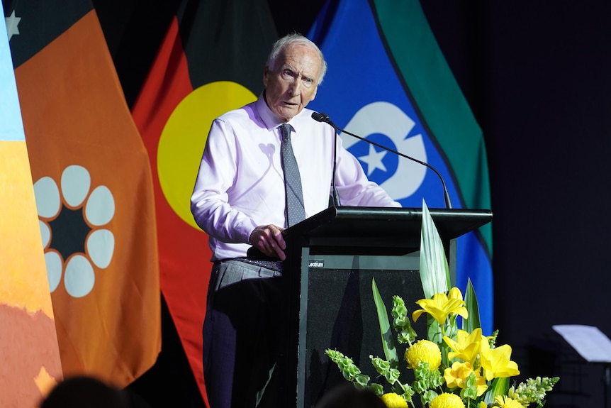 A white man, gray hair, pink button up shirt, gray tie, standing at podium, mid-speech, three flags behind him.