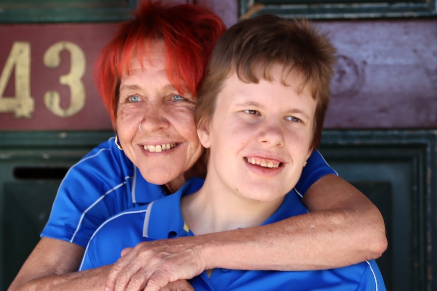 A woman with red hair hugging a teenage girl with a blue shirt in front of a red door with a number.