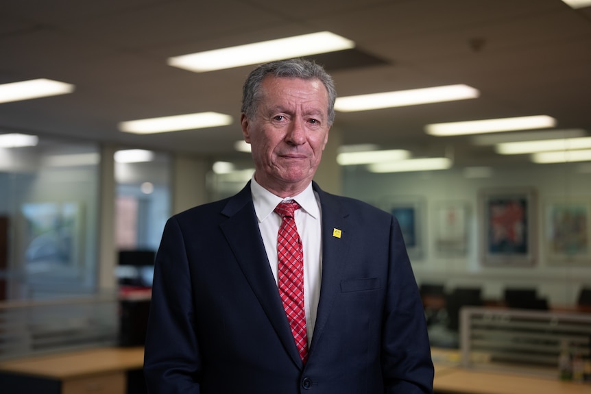 A man in a suit and tie stands in an office, looking into camera with a serious expression.