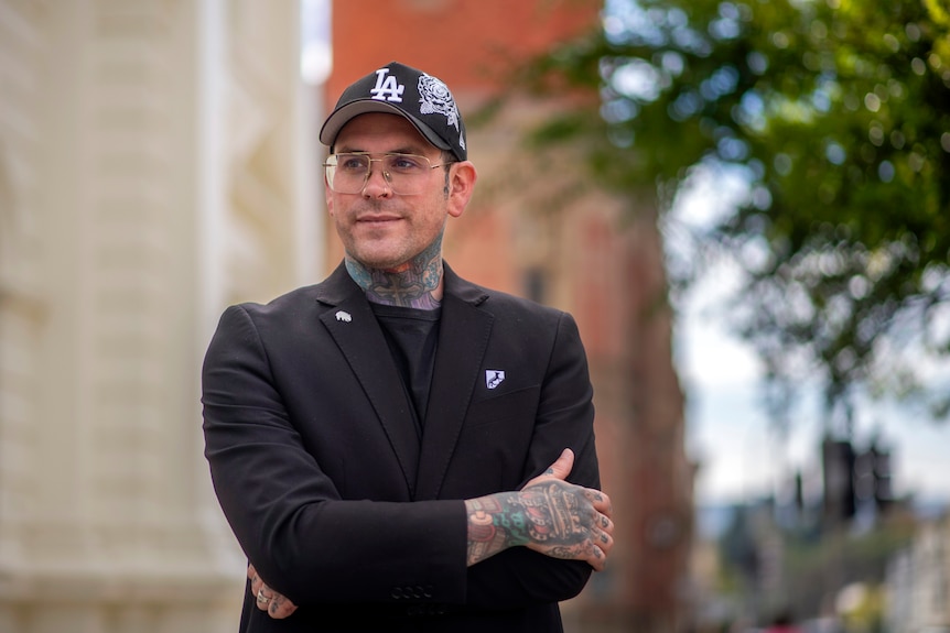 A man dressed in a black with blue neck tattoos smiles with soft white and red buildings and trees in the background.