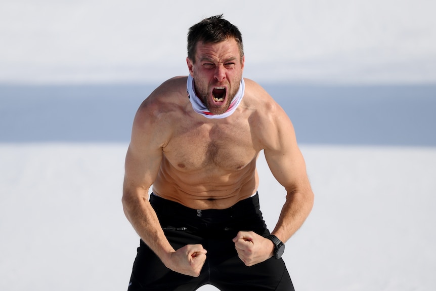 A man with short black hair flexes his muscles while topless in the snow and roars