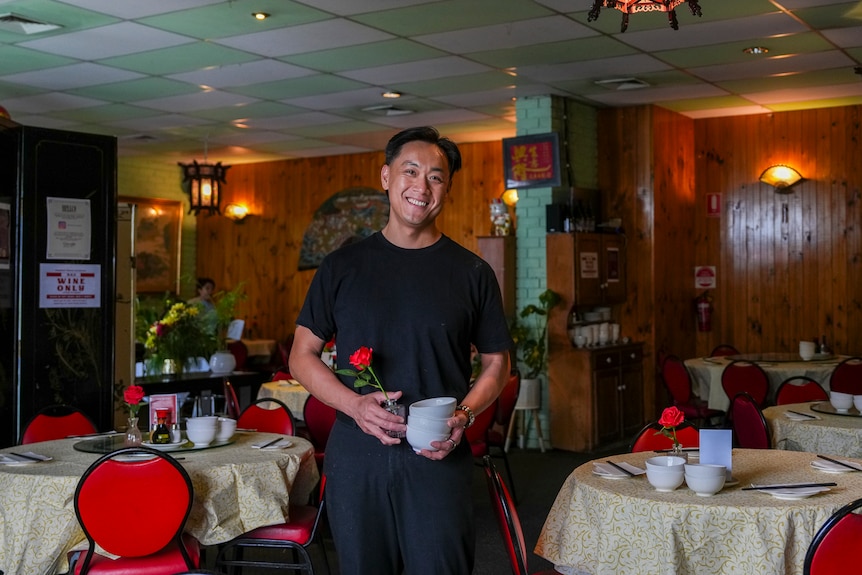 A man wearing black T-shirt standing holding a small flower vase and a stack of bowls inside a restaurant.
