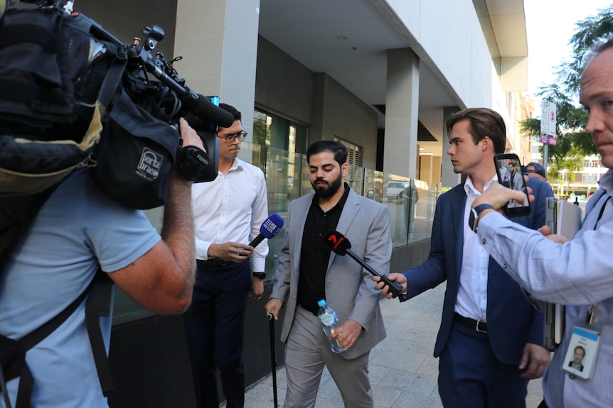 A man walks with a cane being hounded by media. 