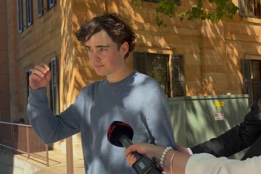 A young, dark-haired man in jumper walks past reporters attempting to question him as he leaves a courthouse.