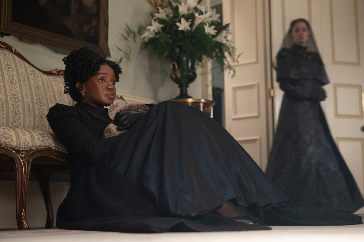 A woman in black period dress sits on the floor by a vintage sofa, appearing somber, with a bouquet of white lilies nearby; another woman in dark attire stands in the background by a door, suggesting a formal or mourning setting.