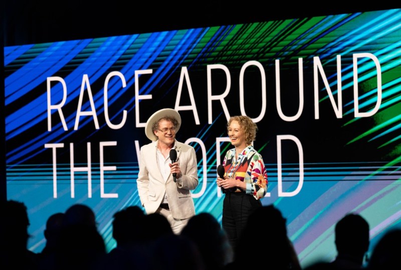 Two presenters speaking on stage at an event titled "Race Around the World," with a vibrant, colourful backdrop. The audience is partially visible, engaged in the presentation.