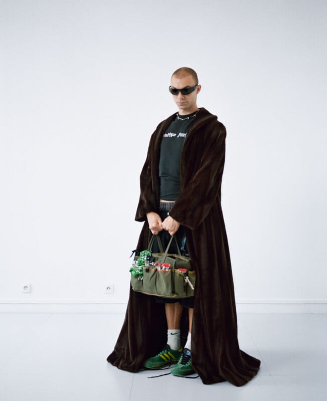 A person wearing dark sunglasses, a long brown robe, a black shirt, and green sneakers stands in a white room, holding a green bag filled with various items and groceries.