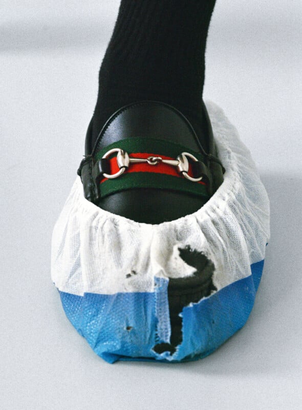 A black loafer with a red and gold detail is worn over a black sock and covered by a disposable blue and white shoe cover that is torn at the front.