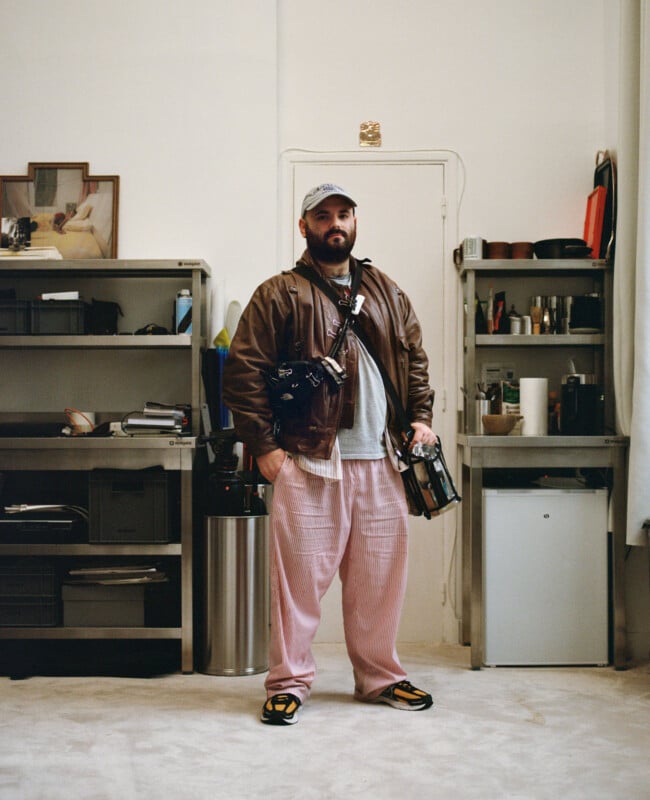 A bearded man stands indoors between two shelves, wearing a brown leather jacket, pink pants, a baseball cap, and colorful sneakers. He carries two bags and holds a camera, facing the camera in a neutral pose.