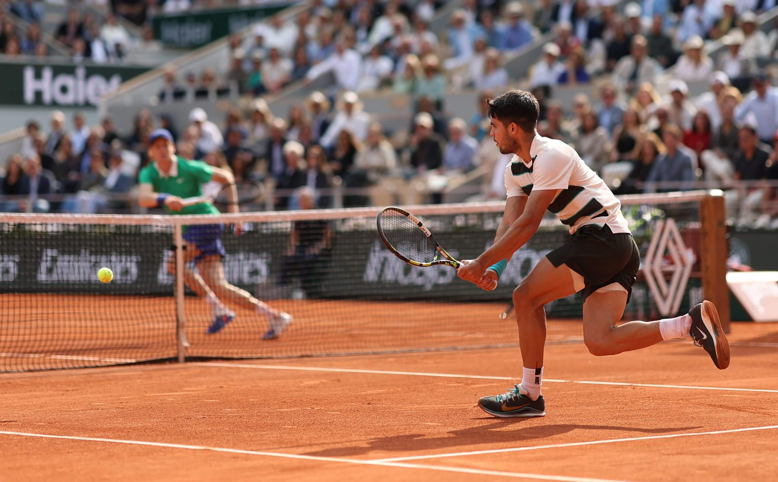 Carlos Alcaraz hits a backhand on a clay court with Jannik Sinner in the background on the other side of the net.