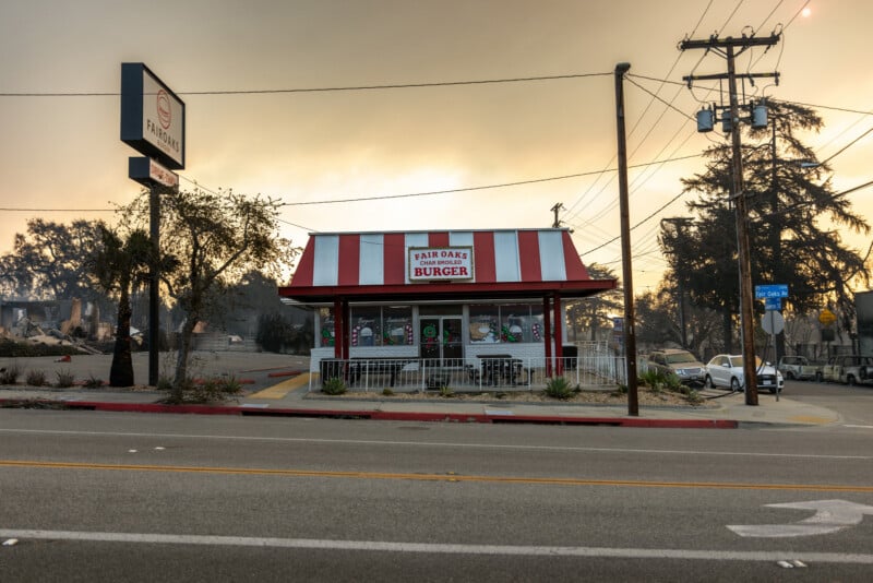 A small diner with a red and white striped roof and “Fair Oaks Burger” sign stands on a street corner. Smoke fills the sky, and burned trees and damaged buildings are visible in the background.