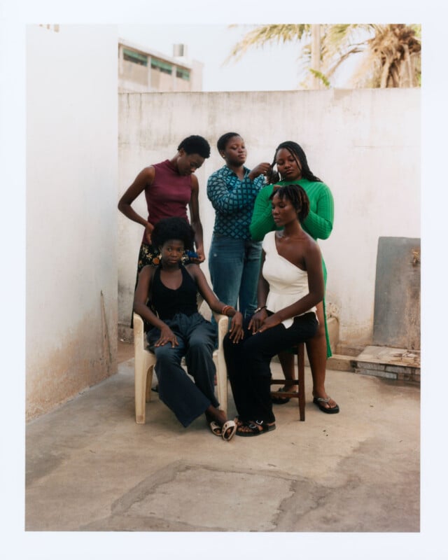 Five women are gathered in an outdoor, walled space. Two are seated on chairs in front, while three stand behind them. One standing woman is braiding the hair of one seated woman. Palm trees and buildings are visible in the background.