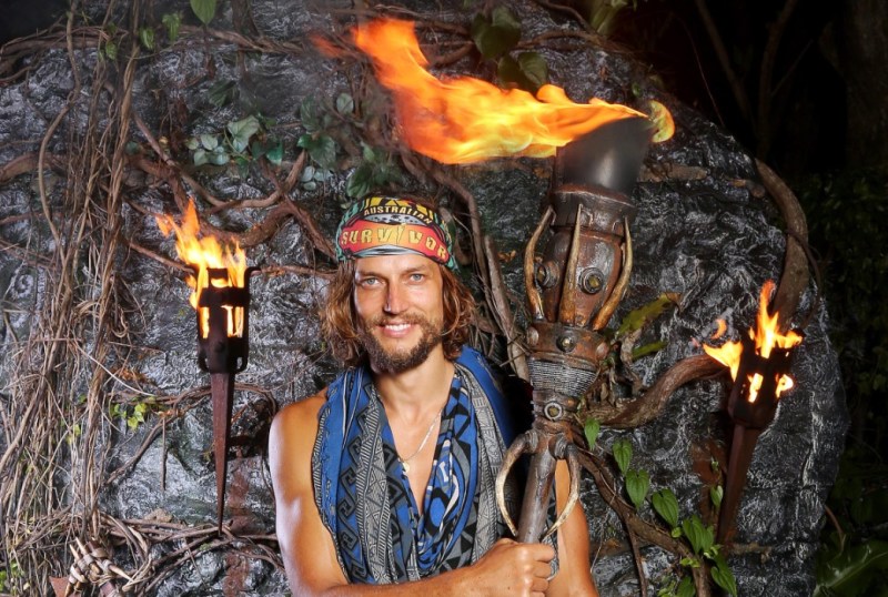 Contestant from Australian Survivor holding a flaming torch, smiling confidently in front of a rocky backdrop adorned with vines and foliage. The image captures the essence of adventure and competition in the reality TV series.
