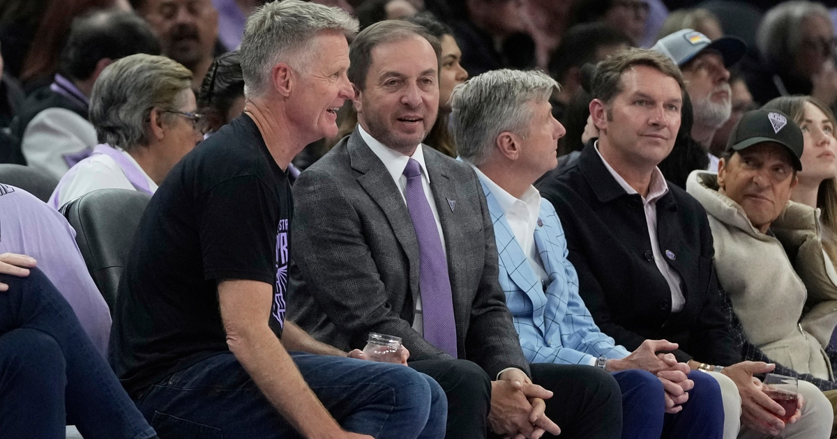 Five men sit closely together in an audience, engaged in conversation, with two holding drinks and wearing casual to business-casual attire.