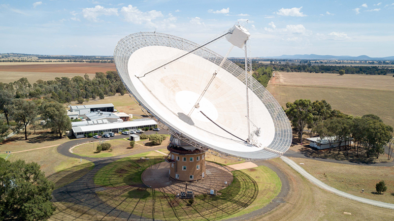 A drone view of a large white dish-shaped radio telescope held up by a terracotta cylindrical building covered in windows. The telescope is shown in daytime surrounded by green fields and trees.