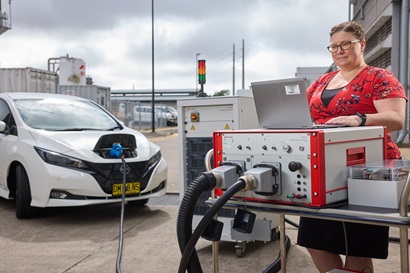 A woman in a red floral shirt operates a laptop connected to electric vehicle charging equipment, with a white EV charging nearby.