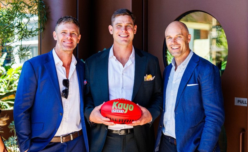 Three men pose together in smart attire, holding a Kayo Sports AFL football, set against a stylish backdrop with greenery. The image captures a moment of camaraderie and excitement, likely related to Australian Rules Football.