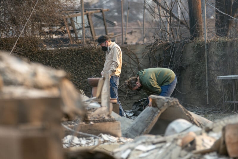 Two people wearing masks sift through the ashes and debris of a burned structure, surrounded by charred remains and damaged objects, likely after a wildfire.