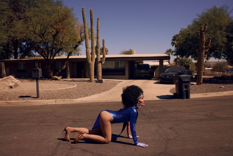 A person in a blue bodysuit, high heels, gloves, and an afro wig crawls on hands and knees on a suburban street, with a house, cacti, and parked cars in the background under a clear sky.