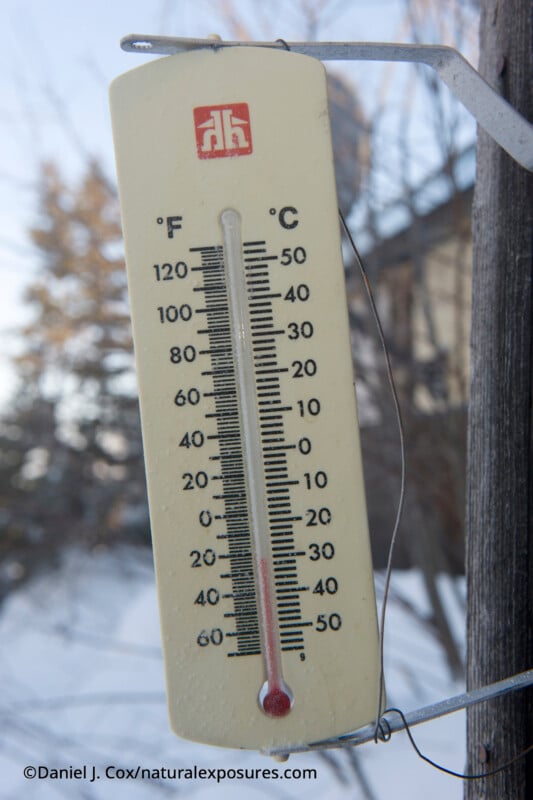 A close-up of an outdoor thermometer attached to a wooden post shows a temperature below 0°F or -20°C, with snow-covered ground and trees blurred in the background.