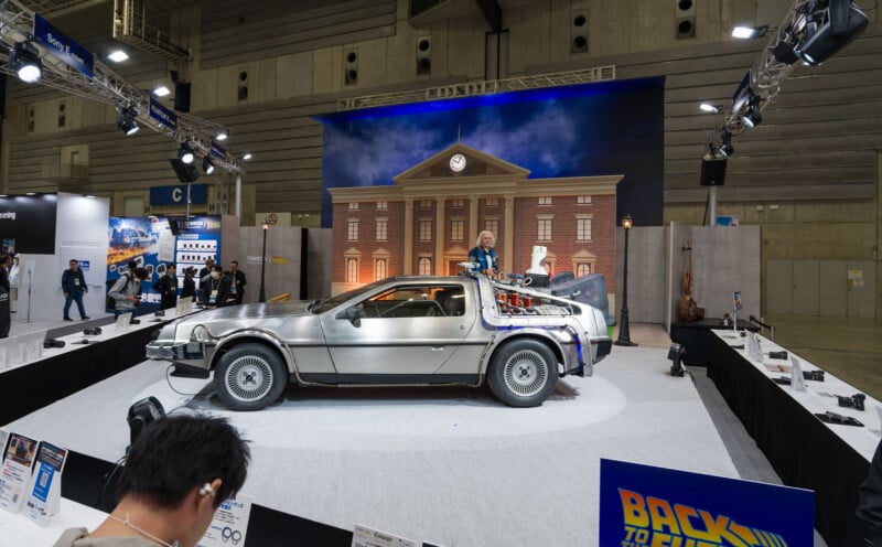 A silver DeLorean car from "Back to the Future" is displayed indoors on a platform, with a clock tower backdrop and a person standing through the roof. Exhibition booths and attendees are visible around the display.