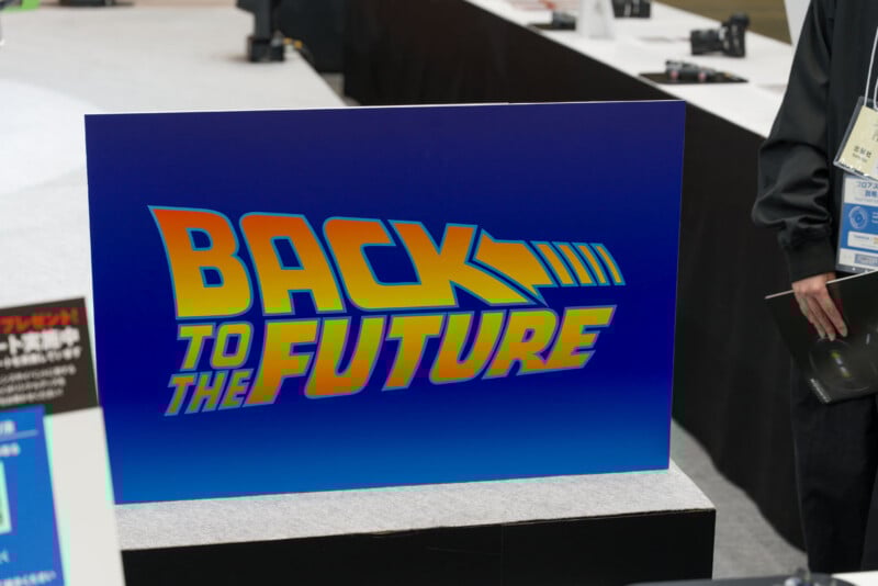 A large sign displays the iconic “Back to the Future” logo in yellow and orange gradient letters on a blue background at an indoor event or exhibition.