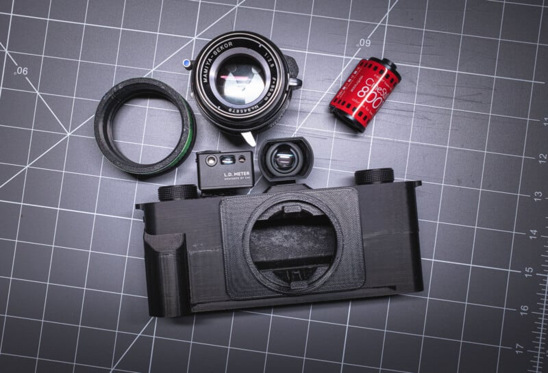 A disassembled film camera is laid out on a gridded cutting mat, showing a lens, a light meter, a viewfinder, a lens adapter ring, a 35mm film canister, and the main camera body.